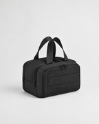 A black nylon toiletry bag with dual top handles, a zippered main compartment, and a front zippered pocket, shown on a light gray studio background.